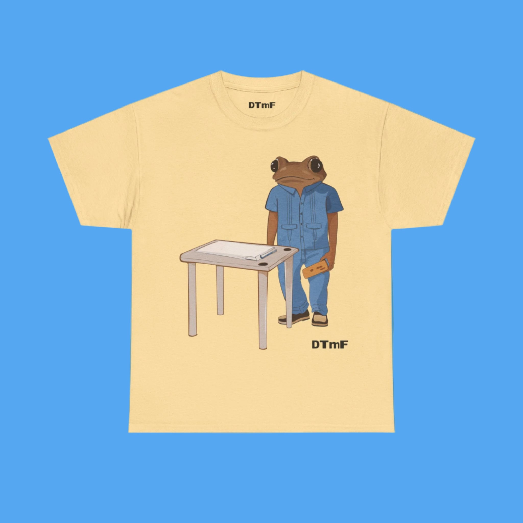 Beige t-shirt with a graphic of a bear in a blue shirt standing next to a table on a blue background.
