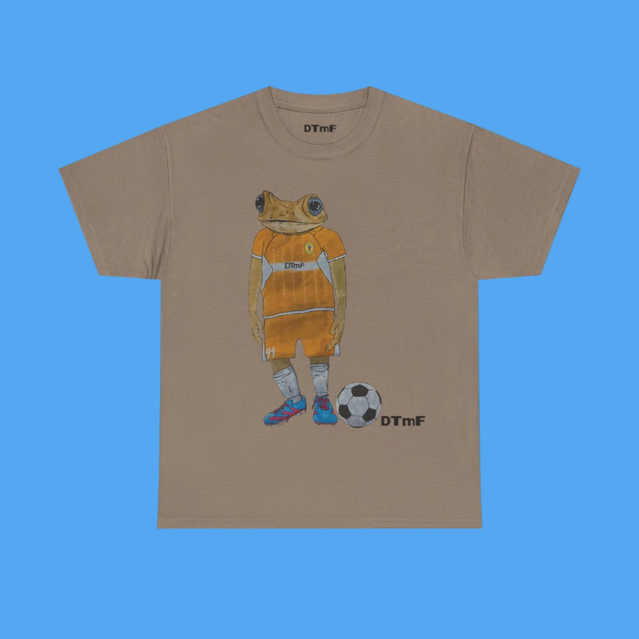 Brown t-shirt with a graphic of a frog in an orange outfit playing soccer on a blue background